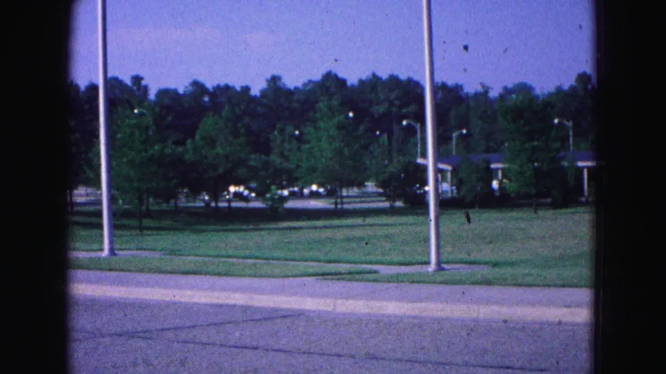Williamsburg-area roadside view from a 1964 8mm home movie, with grass, flagpoles, trees, and a low modern building.