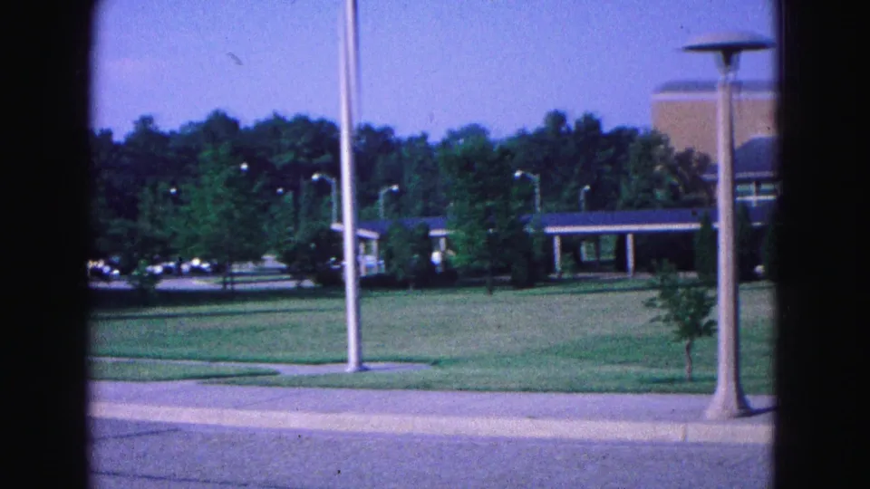 1964 Williamsburg roadside travel shot with lawn, tall poles, trees, and a low building glimpsed from a moving car perspective.