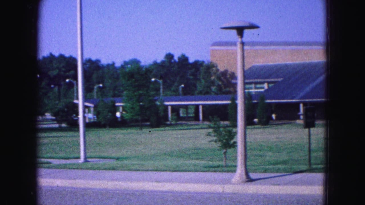 1964 Williamsburg-area 8mm frame showing a low blue-roofed building across grass with young trees, signposts, sidewalk curb, and tall lampposts.