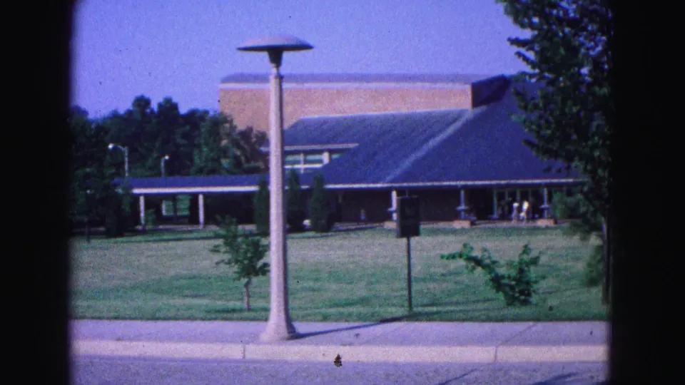 1964 Williamsburg-area exterior view of a blue-roofed building with lawn, trees, lampposts, and distant pedestrians near the entrance.