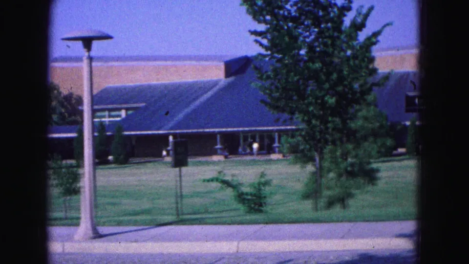 1964 Williamsburg-area frame with a low blue-roofed building, central lamppost, leafy tree, lawn, sidewalk curb, and distant entrance area.