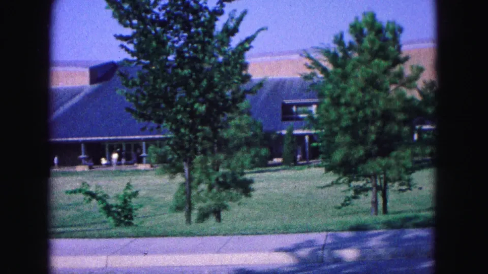 1964 Williamsburg-area 8mm frame showing trees in the foreground, a blue-roofed building, lawn, sign, and distant entrance activity.
