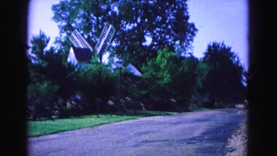 A white windmill appears among dense trees and greenery near a quiet Williamsburg street in the 1964 8mm footage.
