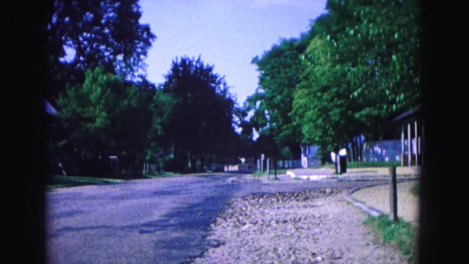 End frame of the 1964 Williamsburg home movie showing a quiet road, mature trees, roadside stones, sidewalk, and low buildings at the right edge.