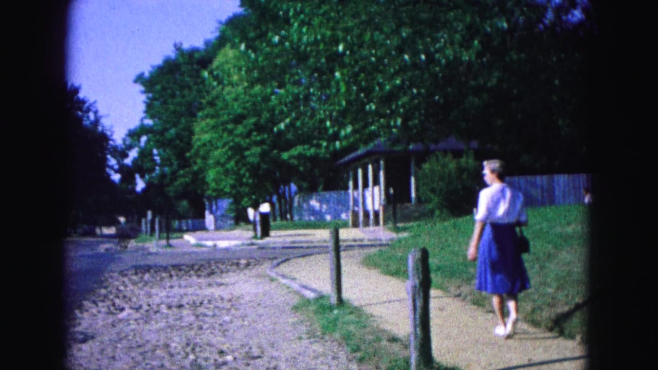 Woman in a white blouse and blue skirt walks along a path beside trees and posts.