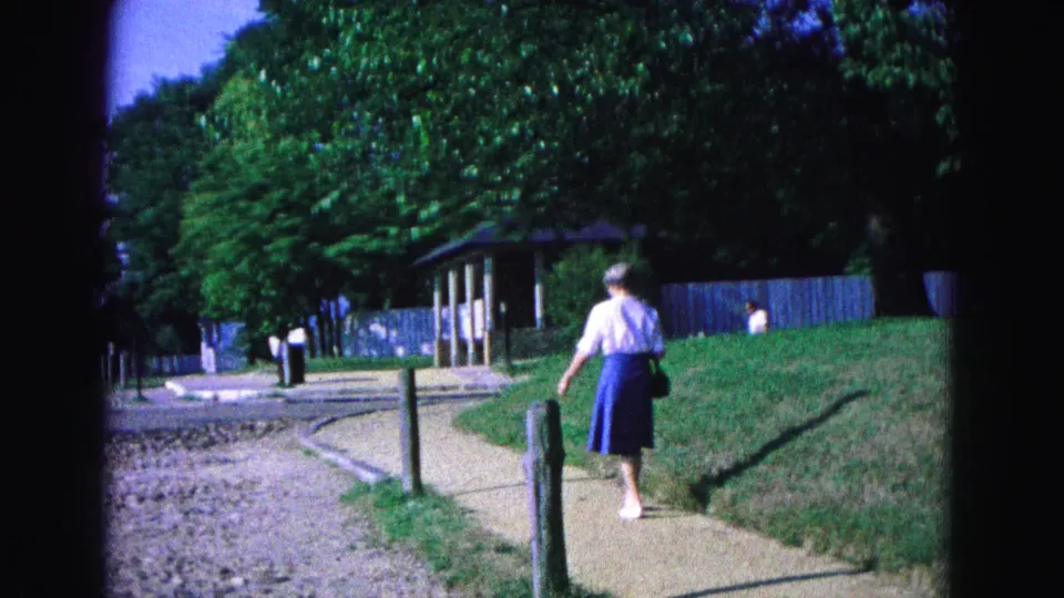 A woman in a white blouse and blue skirt walks away along a Williamsburg path in this 1964 8mm home-movie frame.