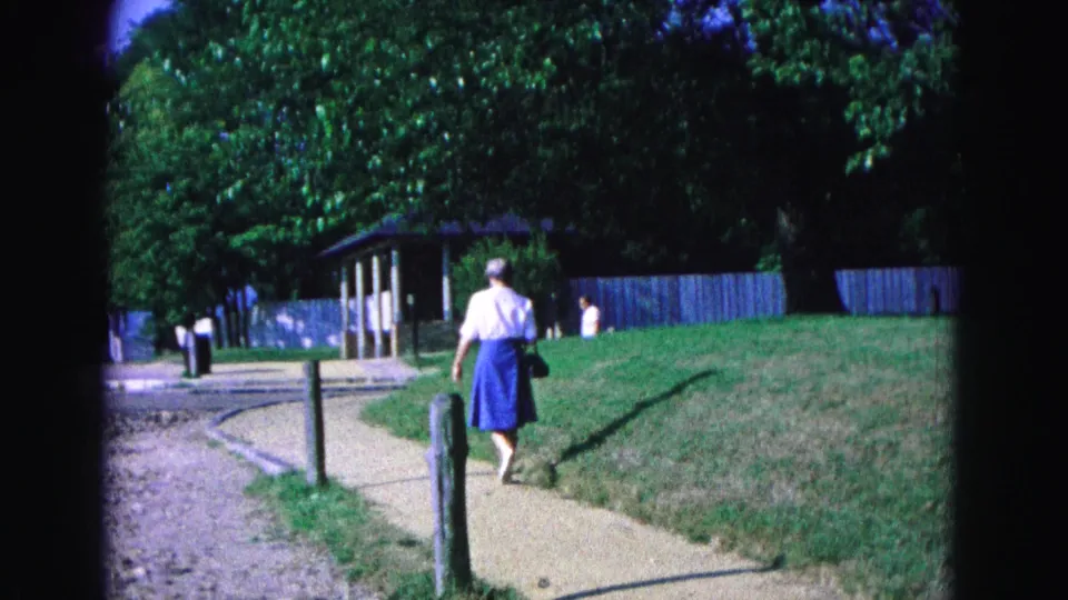 The 1964 Williamsburg home-movie view shows the woman farther along the path, with trees, grass, posts, and distant pedestrians ahead.