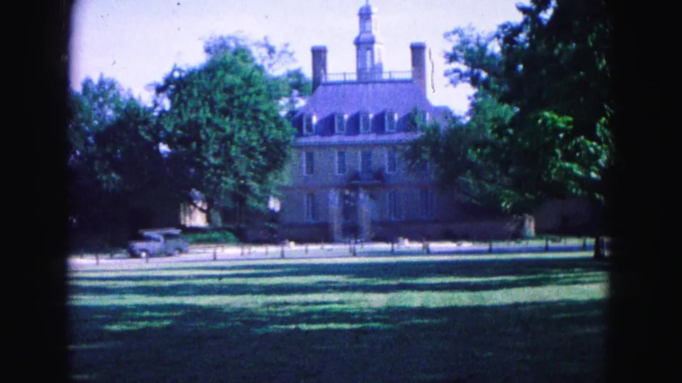 1964 Williamsburg home-movie still showing the brick building and cupola partly framed by trees with a pickup on the road.