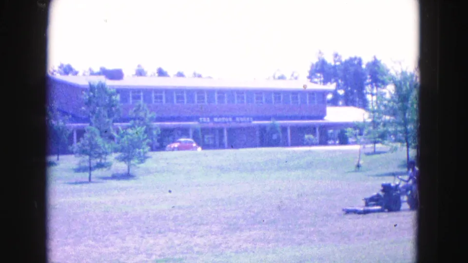 1964 Williamsburg frame showing a low modern building, a red car, open lawn, and a distant person operating a mower.