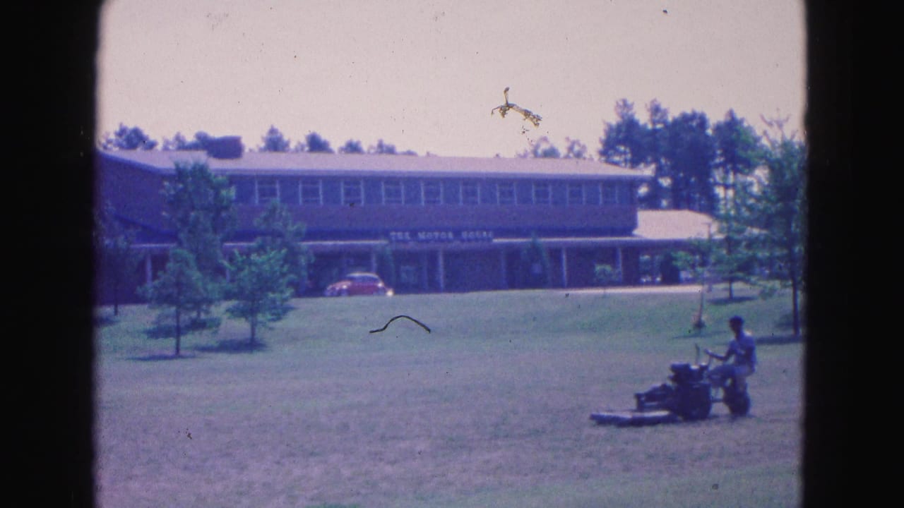 Man rides a lawn mower across grass in front of a large two-story building.