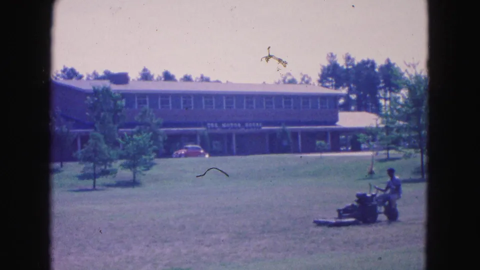 1964 Williamsburg home-movie frame showing a man on a lawn mower before a broad mid-century building and trees.