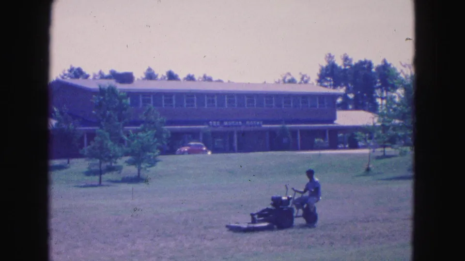 A man continues mowing a sunlit Williamsburg lawn in front of a signed mid-century building in 1964 footage.