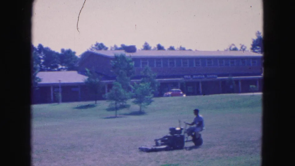 1964 Williamsburg frame with a man mowing below a long flat-roof building, trees, and a parked red car.