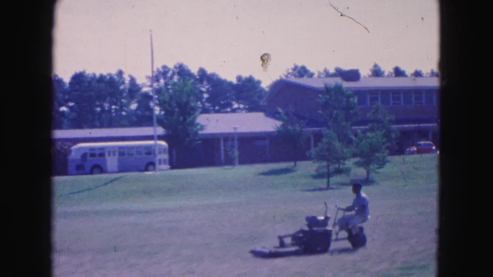 A 1964 Williamsburg frame shows a man mowing at right with a bus, trees, and a large building behind him.