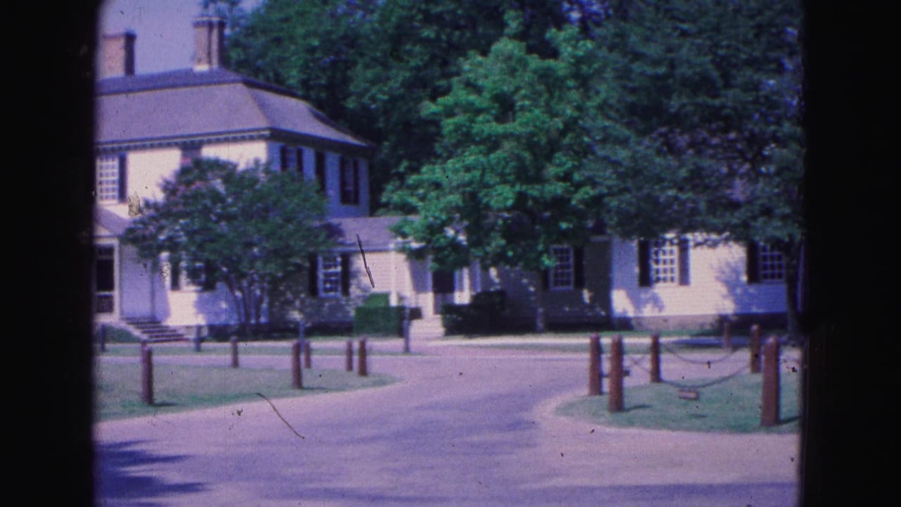 Historic white building with dark shutters beside paths, lawns, trees, and posts.