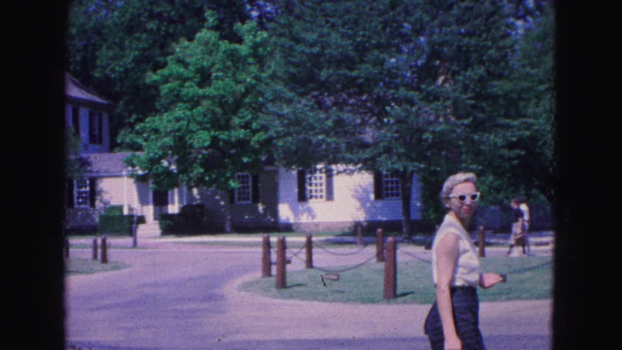 Woman in sunglasses walks near a path with historic buildings and trees behind her.