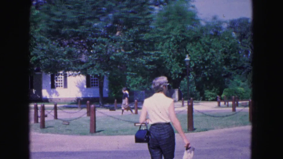 The walking visitor is seen from behind on Williamsburg grounds, carrying a handbag beside paths, lawns, trees, and shuttered buildings.