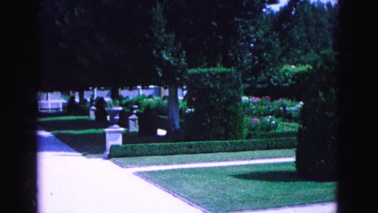 Formal garden path bordered by hedges, lawn, trees, stone posts, and a white fence.