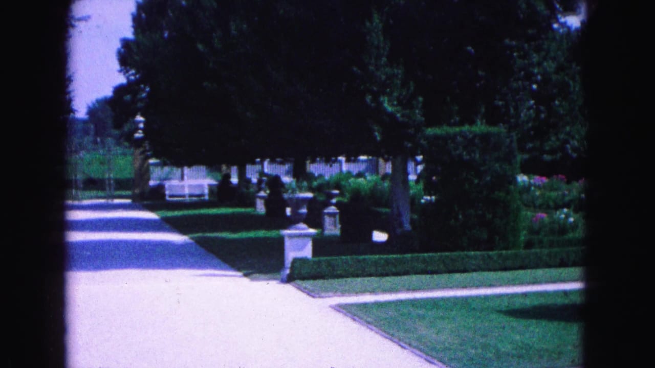 Garden walkway with hedges, lawn, trees, stone ornaments, and a white fence beyond.