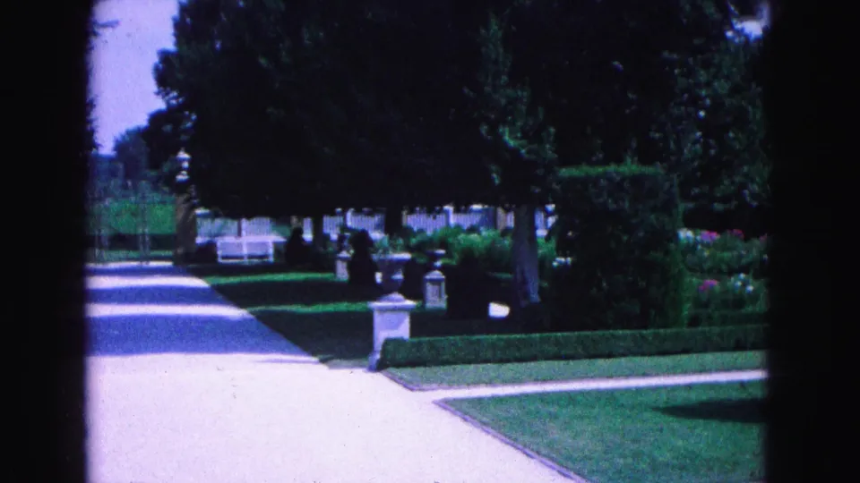 The pan continues across a Williamsburg garden path lined by manicured hedges, stone ornaments, lawn, trees, and a distant white fence.