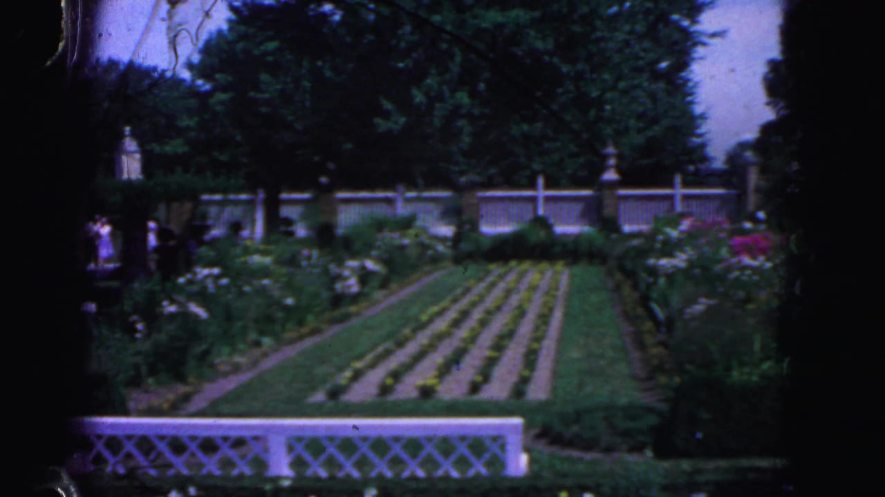 Formal garden beds behind a white lattice fence with trees and distant visitors.