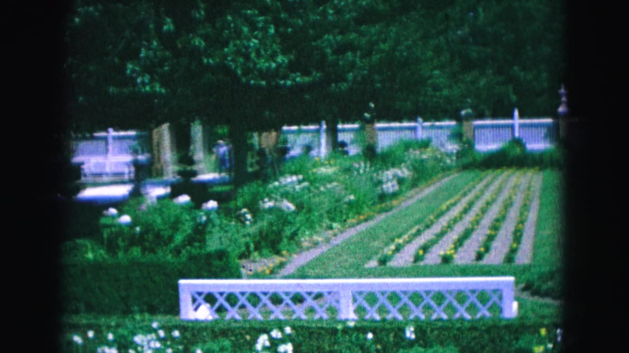 Formal garden with white lattice fence, flower beds, trees, and distant visitors.