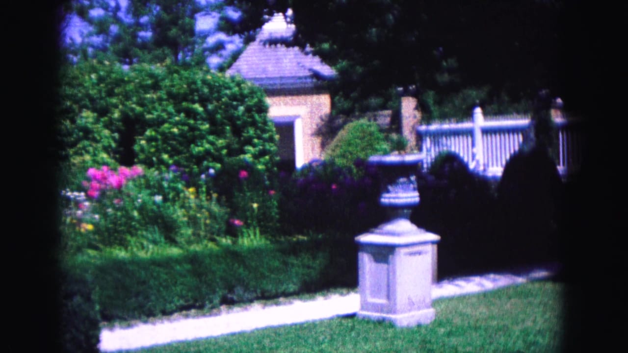 Garden view with pink flowers, hedges, a stone planter, white fence and lawn.