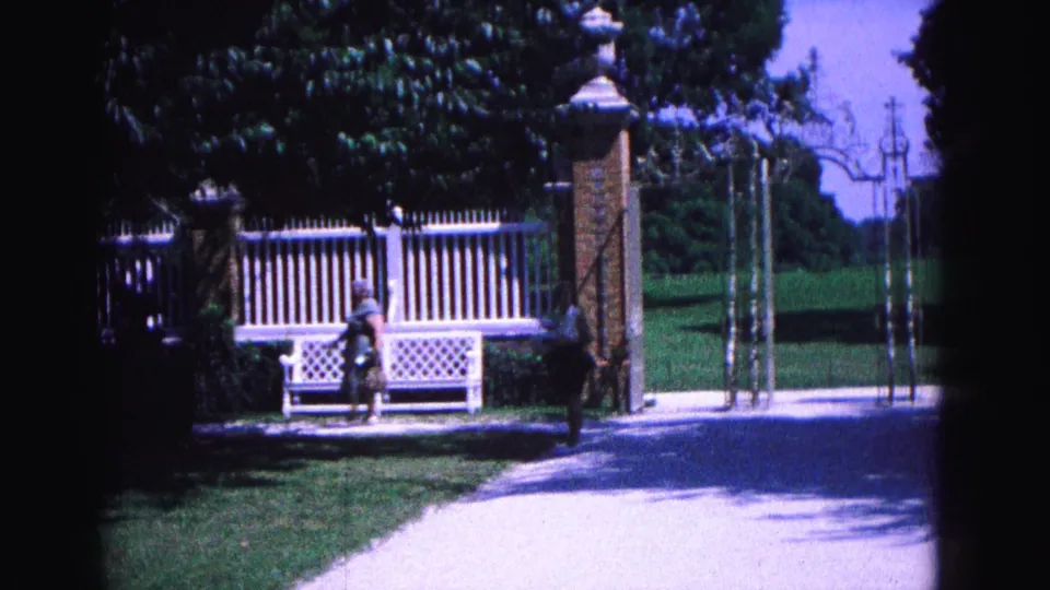 In 1964 Williamsburg footage, a woman in a light dress walks beside a white lattice bench near brick gate pillars and a fence.