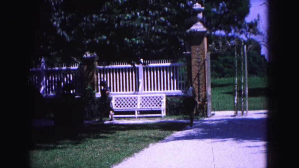 Williamsburg garden scene with a woman walking past a white lattice bench, brick gate pillars, fence, lawn and trees.