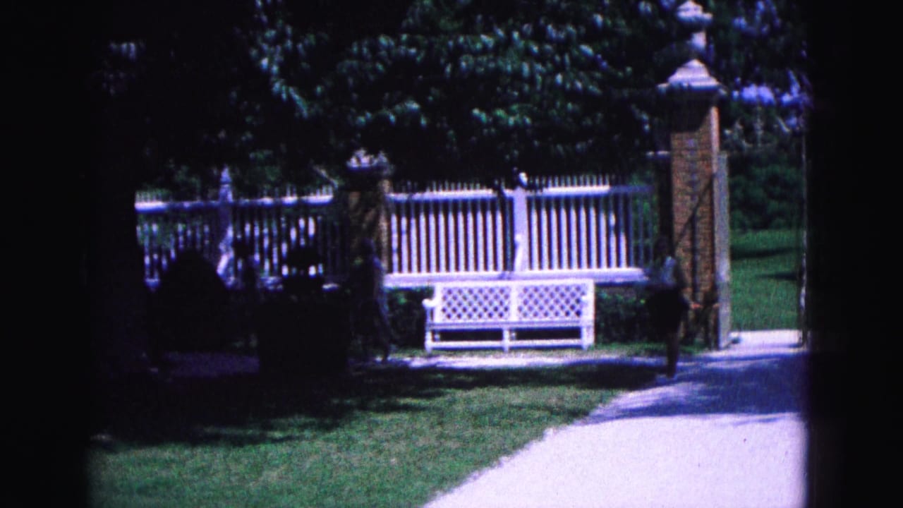 White picket fence, lattice bench, brick gate posts, lawn, trees, and a paved path.