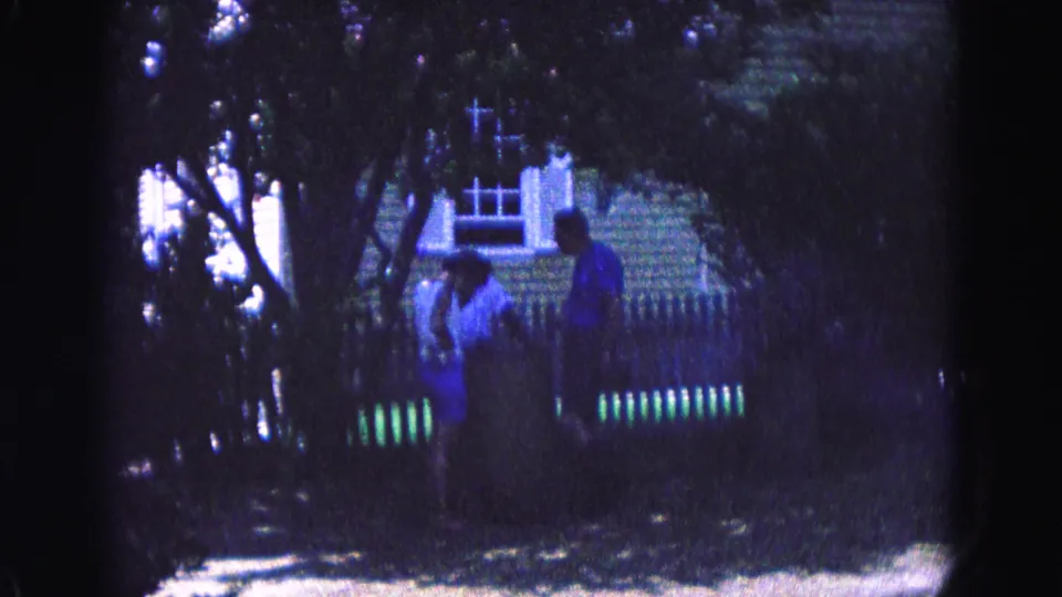 1964 Williamsburg-area home-movie frame with two distant people walking under trees near a fence, shrubs, and a building facade.