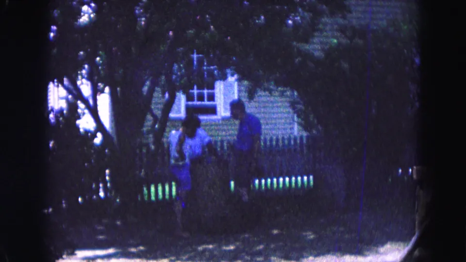 1964 Williamsburg-area 8mm frame showing two distant people under heavy tree shade, with a fence, shrubs, and building behind them.
