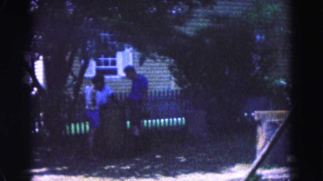 Two people stand near a white fence in a leafy yard.