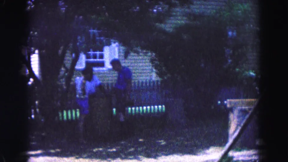 Two people appear near a white picket fence in a leafy Williamsburg yard, filmed in soft 1960s 8mm color.