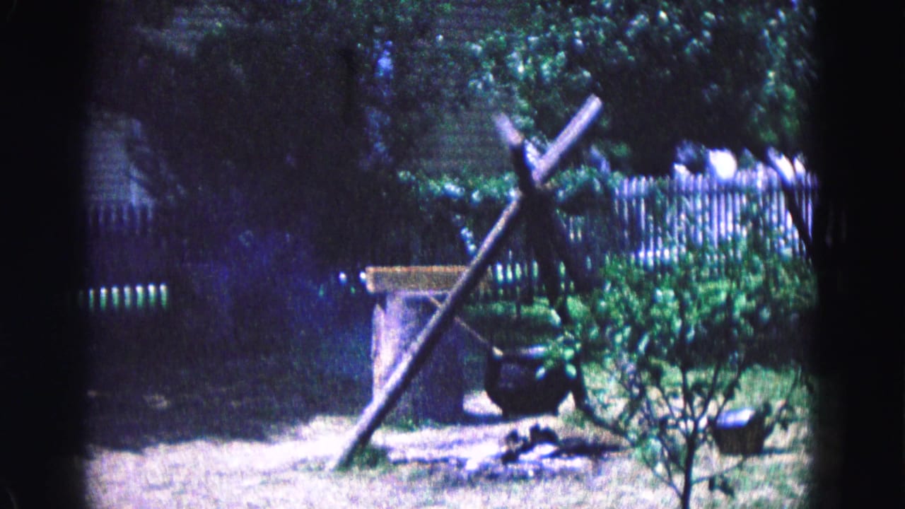 Wooden tripod with a hanging cauldron beside shrubs and fencing.