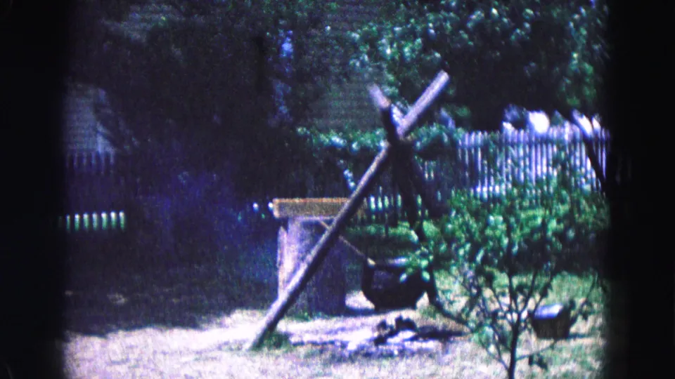 A rustic wooden tripod and hanging cauldron stand near shrubs and a white fence in Williamsburg, Virginia.