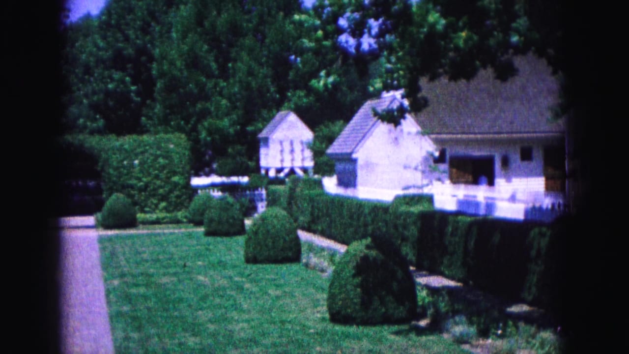 Manicured lawn with hedges, walkway, white fence, and white houses beyond trees.