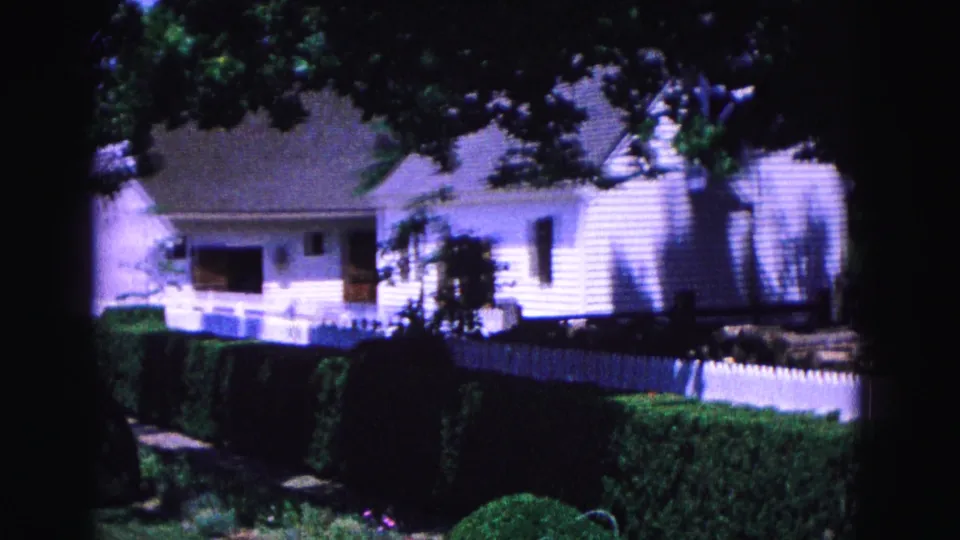 A soft 8mm frame of a white house behind picket fencing and hedges, with tree shadows across the scene in Williamsburg.