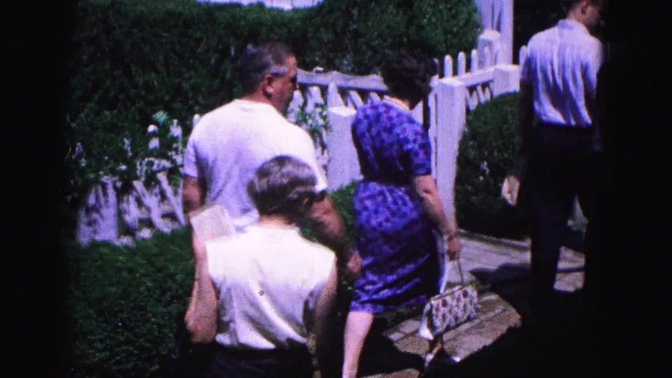Adults in summer clothing walk away along a Williamsburg path bordered by hedges, a white fence, and residential buildings.