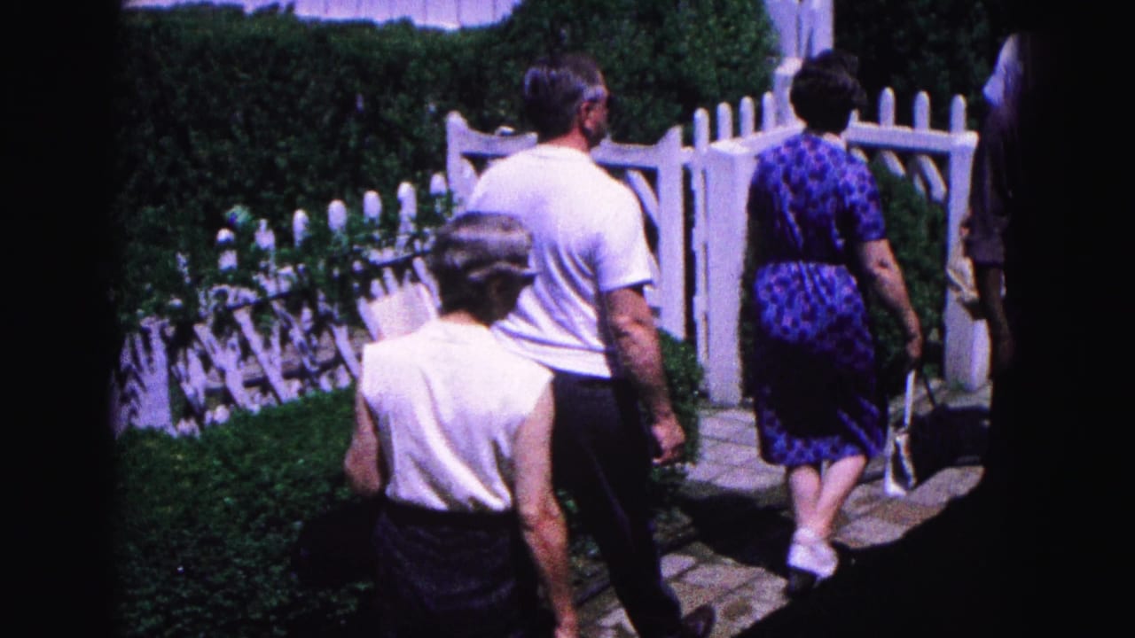 Adults walk along a garden path beside a white picket fence and hedges.