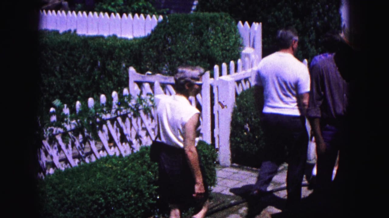 Adults in 1964 Williamsburg footage move through a garden entrance area bordered by white fencing, hedges, and bright sunlight.