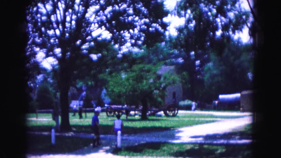 1964 Williamsburg home-movie frame showing visitors on shaded park grounds, with trees, lawn, a light road, cannons, and a distant structure.