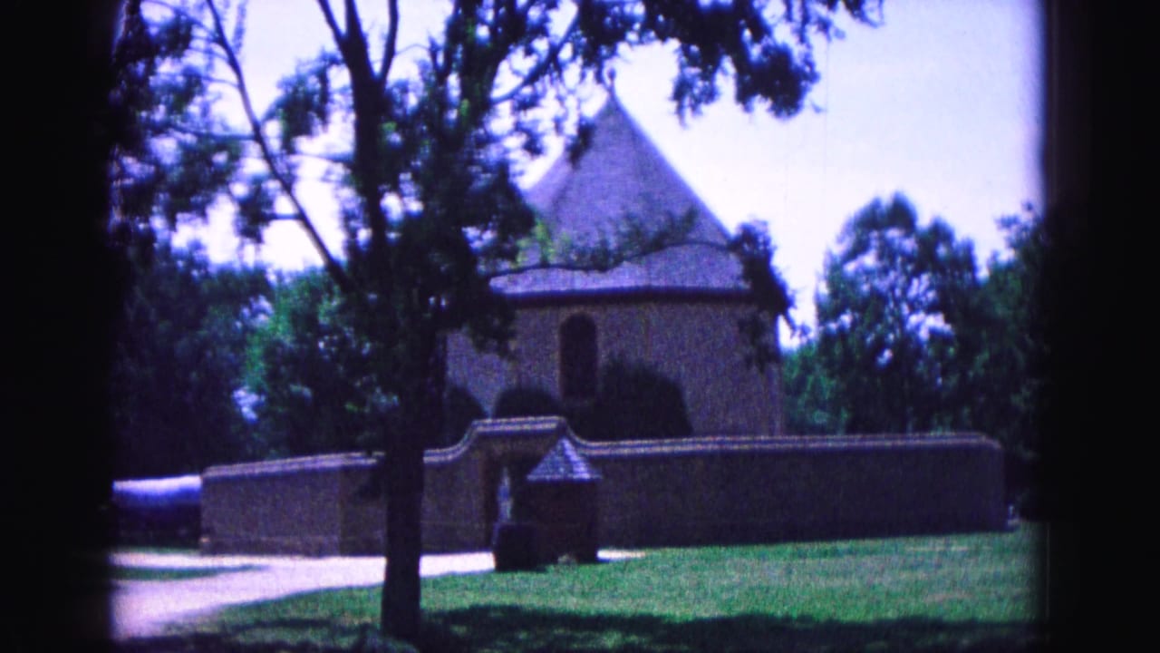 Historic building behind a low wall, with a large tree, lawn, and road in front.