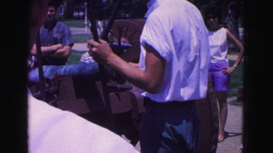 1964 Williamsburg home-movie frame showing a man in a white shirt working at the cannon with visitors watching nearby.