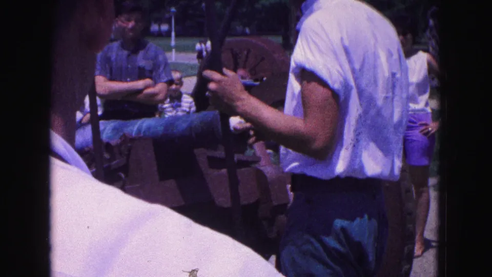 1964 Williamsburg frame showing the cannon demonstration from a close visitor perspective, with adults watching in summer clothes.