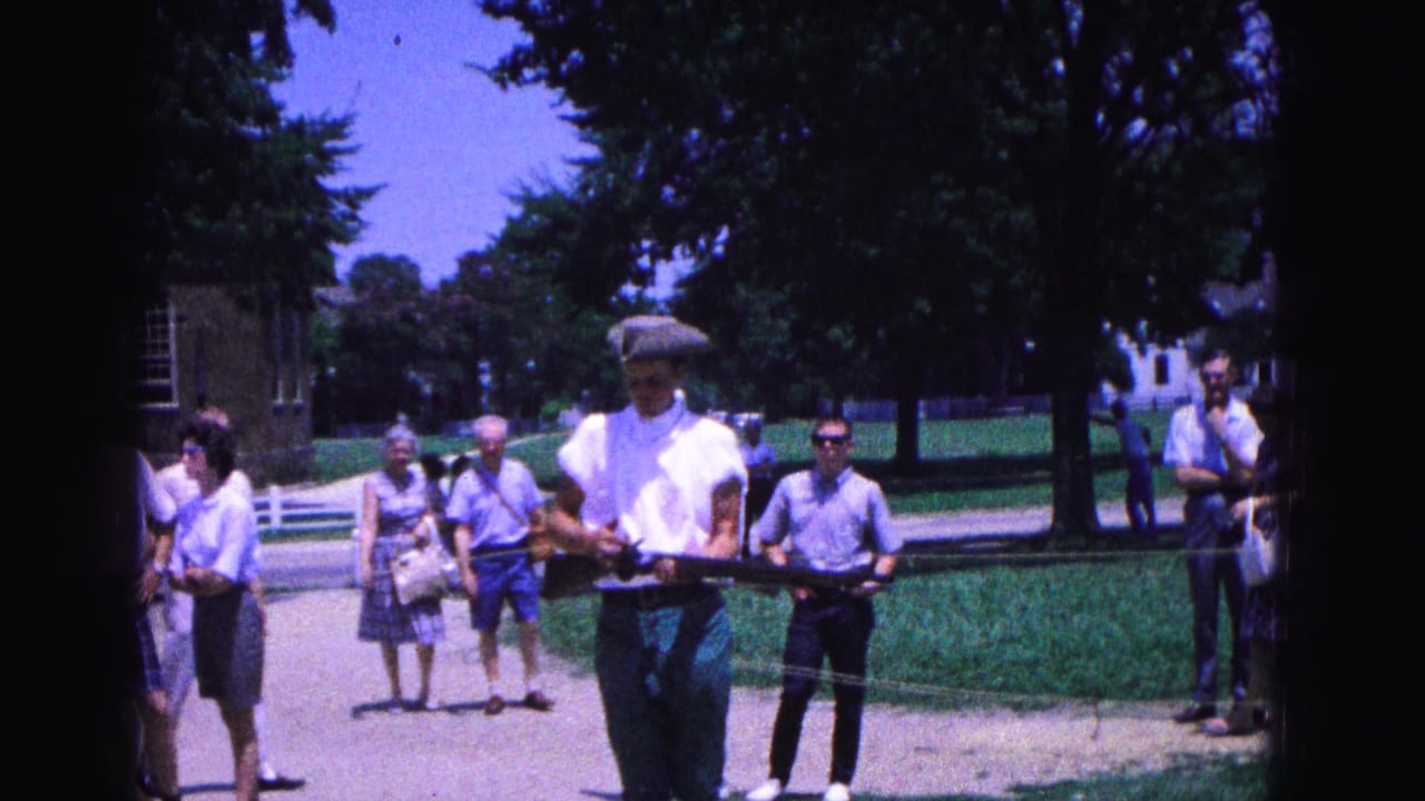 In 1964 Williamsburg, Virginia, a costumed marcher with a replica musket moves forward as visitors watch from a sunny walkway.