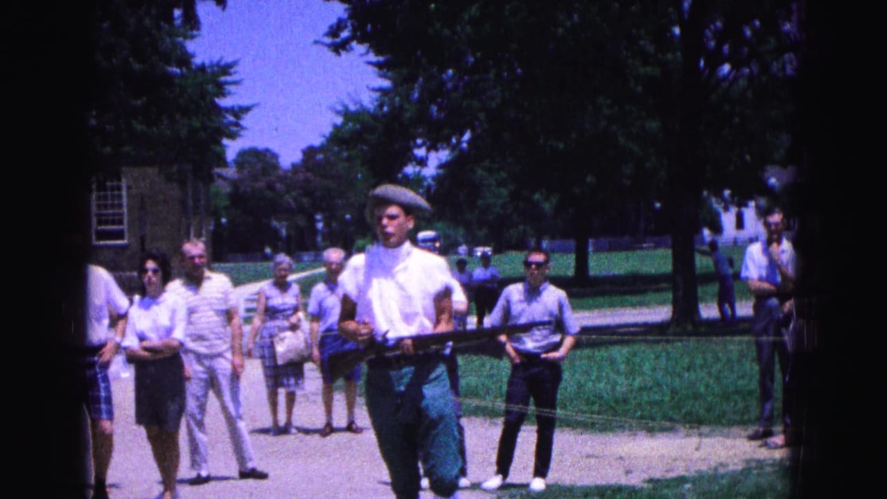 A Williamsburg reenactment participant in white shirt and cap carries a replica musket with spectators and a historic-looking building behind him.