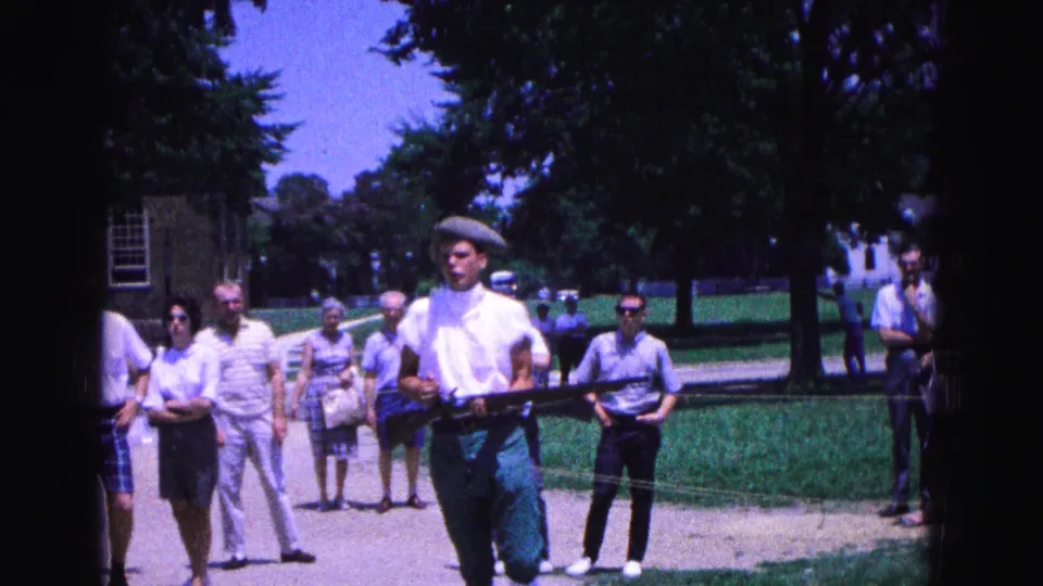 A Williamsburg reenactment participant in white shirt and cap carries a replica musket with spectators and a historic-looking building behind him.