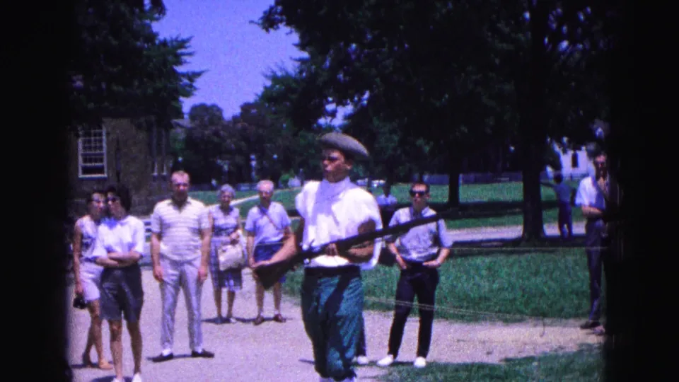 The 1964 home-movie frame shows the reenactor moving across the foreground with casually dressed visitors watching in Williamsburg.