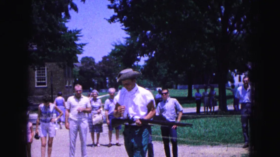 A costumed Williamsburg reenactor carries a replica musket across the frame as visitors stand near a rope line and shaded lawn.